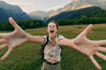 Naklejka premium Happy woman with backpack stretching arms towards camera in green meadow with mountains in background. Outdoor lifestyle, adventure, travel, and freedom concept.