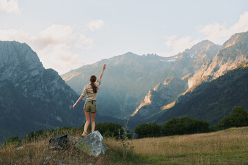 Naklejka premium Woman outdoors on a rock in a meadow, arms raised in a carefree pose, majestic mountains in the background, sunlit scene, natural colors, lifestyle moment