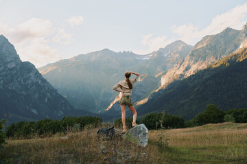 Naklejka premium Young woman stands on a rock in a sunlit valley, arms raised in triumph as distant mountain peaks glow under a clear blue sky, wearing beige tank top and light shorts, embodying freedom