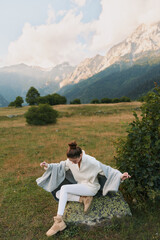 Naklejka premium Woman outdoors in a meadow near mountains, wearing a light sweater and white pants, sitting on a rock with a cozy cape, enjoying a relaxed sunlit moment.