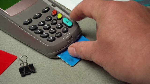 Hand inserting blue credit card into card reader while pressing buttons, with paper and office supplies visible on the table