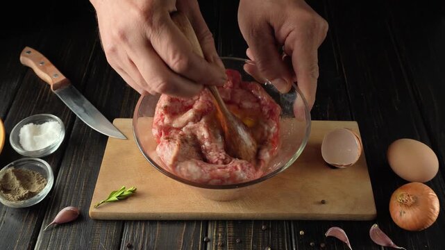 Hands mixing ground meat and egg in a glass bowl on a wooden cutting board with spices and vegetables arranged around in a kitchen setting