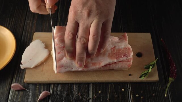 Hand slicing pork fat on a wooden cutting board with garlic cloves and green herbs visible in a well-lit kitchen setting