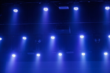 Naklejka premium Cool blue LED stage lights shine through haze from a dark ceiling rig, creating a moody concert or theater background with copy space