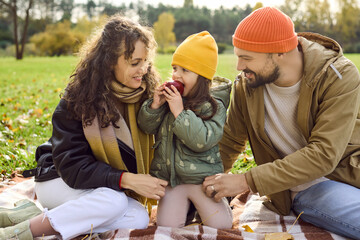 Naklejka premium Family enjoys autumn picnic with happy daughter. Parents relax on a blanket in the park as their child enjoys a red apple, bundled in warm clothing. Happy family time outdoors in sunlight.
