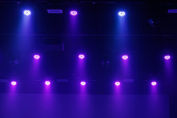 Naklejka premium Purple and blue LED stage lights shine through haze from a dark ceiling rig, forming a moody concert background with copy space