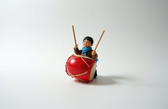 Figurine of child plays bright red drum with sticks. This detailed tiny musician figure wears brown jacket and blue scarf. Small drummer is posed on white background, ready to make some beat.