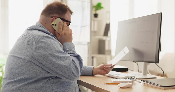 Man on phone about bill and expenses. At home by computer he reviews payment document and sets a budget plan to manage utility charges and invoices. Theme is personal finance and payments.