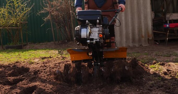 Man operating a motor cultivator to till the earth in a garden plot. Agricultural machinery preparing the soil for spring planting, loosening and aerating the ground for farming.