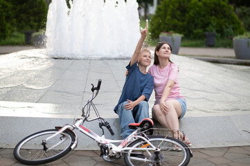 Fototapeta premium Happy mother and son sitting by a city park fountain during a bike ride. The young boy is pointing at something in the sky or distance, while the woman looks on with a smile. Concept of family bondin
