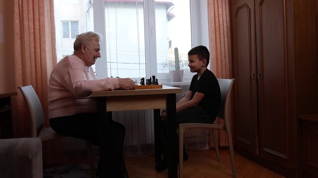 Grandmother playing chess with grandson at the table by the window. A good way to spend time