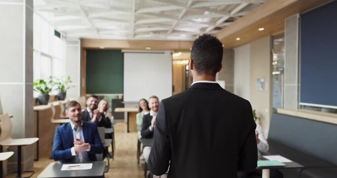 Man Leading Business Meet Training From Back View, Presenter In Suit Addressing Attentive Team In Modern Office, Attendees Clapping And Taking Notes, Flipchart And Laptop Visible,