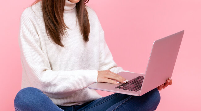 Student in jeans sitting on floor and using silver laptop on her lap for online education or surfing isolated over pink background