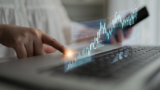 A woman engages with data on her laptop while using a smartphone, showcasing holographic financial graphs and trends in a contemporary workspace. Scalp