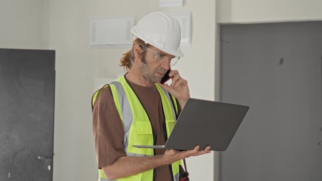 Man in reflective vest holds laptop and phone to ear while checking screen inside building; concentration planning.