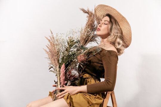Dreamy woman in straw hat leaning on chair with flowers bouquet holding glass vase with closed eyes and calm face isolated on white background