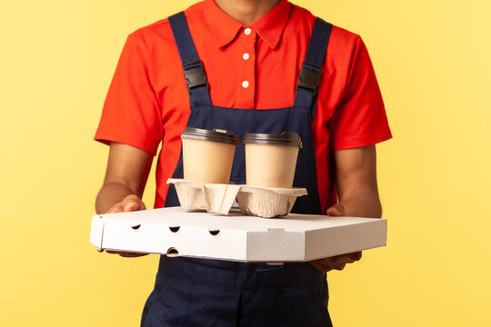 Delivery service. Friendly professional courier in blue overalls holding coffee and pizza box, wearing, offering drinks and food, looking at camera. Indoor studio shot isolated on yellow background.