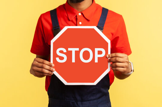 Portrait of handyman wearing blue overalls red hat and t-shirt holding red stop sign, prohibitions, looking at camera with serious expression. Indoor studio shot isolated on yellow background.