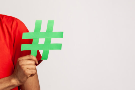 Subscribe to popular blog! Bearded man with dreadlocks wearing red casual style T-shirt, holding social media hashtag symbol and smiling. Indoor studio shot isolated on gray background.