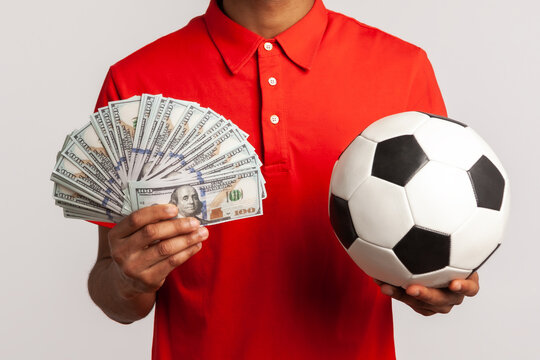 Smiling satisfied man with dreadlocks wearing red casual style T-shirt, holding soccer ball and fan of dollar bills, sports betting, big win. Indoor studio shot isolated on gray background.