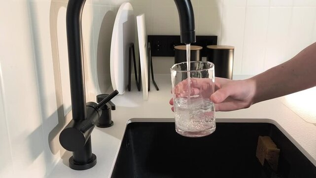 Hand fills glass with filtered water from kitchen tap at home. Clean liquid flows through purification system into transparent container. Healthy lifestyle routine supports hydration and