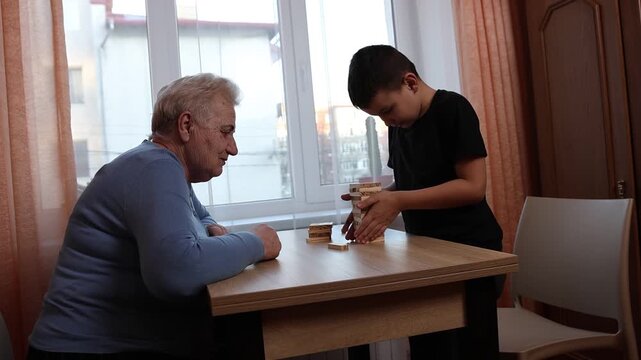 Grandmother and grandson play a tabletop game with wooden blocks, carefully stacking pieces together by the window.