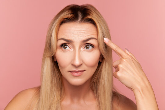 Concerned blonde lady pointing at her forehead skin texture while checking for fine lines and aging signs during beauty routine isolated on pink background