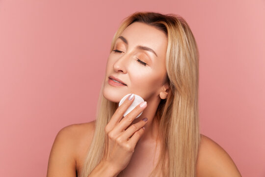 Middle aged woman massaging her chin with soft white cotton pad for deep skin cleaning and aesthetic hydration treatment isolated on pink background