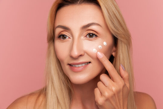 Mid-adult woman with blonde hair pointing to three moisturizing cream dots under her eye during skincare isolated on pink background
