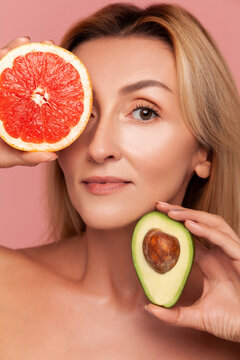 Close up of woman holding grapefruit and avocado halves near eyes, presenting variety of natural vitamins for skin therapy isolated on pink background