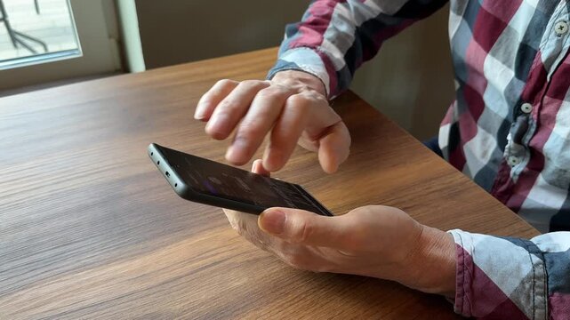 Man using a smartphone at a wooden table. Hands interact with a mobile phone in a bright indoor setting. Digital communication, app browsing, and everyday technology use..