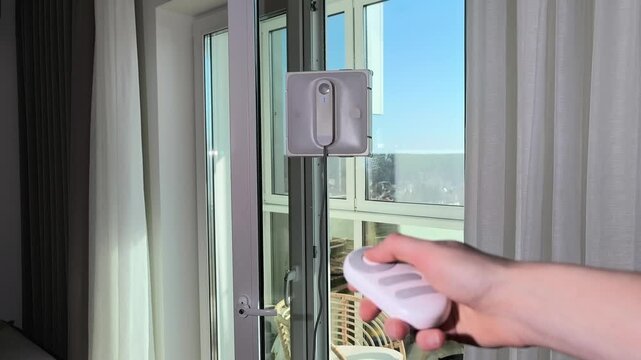 Hand uses remote control to activate robot cleaner on window glass. Device responds to command starting automated washing movement. Spring cleaning routine demonstrates convenient home