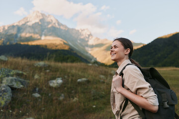 Naklejka premium Woman hiking outdoors with a backpack, smiling as she walks through a sunlit meadow, with distant mountains and lush scenery, showcasing a relaxed lifestyle.