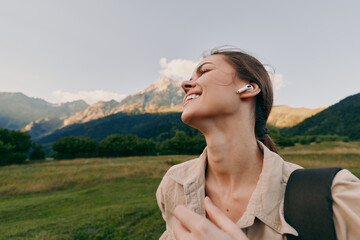 Naklejka premium Smiling woman in a beige jacket outdoors, backpack on shoulder, enjoying nature amid green fields and towering mountains, soft sunlight highlights a relaxed lifestyle moment.