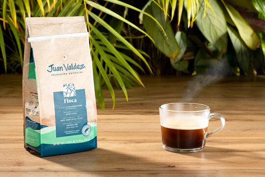 Juan Valdez Finca Colombian whole bean coffee packaging with steaming espresso cup on wooden table and tropical leaves background.