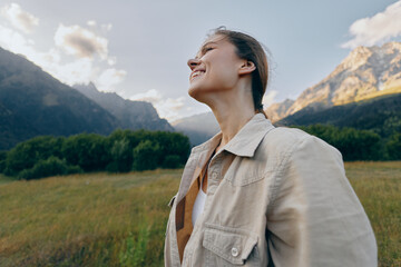Naklejka premium A young woman in a beige jacket stands in a sunlit mountain meadow, tilting her head back with a bright smile, enjoying the fresh air and wide open sky.