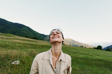 Naklejka premium Young woman with short hair smiles joyfully while standing in a green meadow surrounded by mountains under a clear sky during daytime.