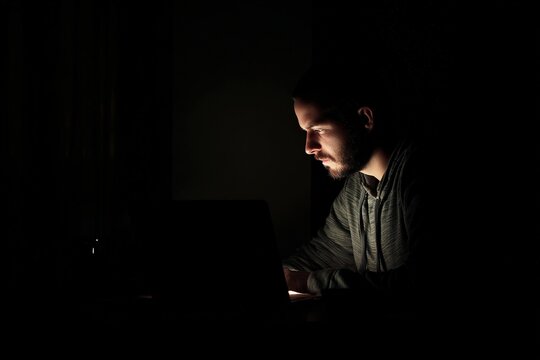 Young bearded man concentrated working on laptop in dark room lit only by screen light at night