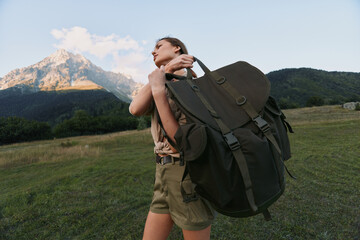 Naklejka premium A woman with a large backpack stands in a meadow with mountains in the background, preparing for a hike. An outdoorsy lifestyle moment emphasizing adventure and travel.