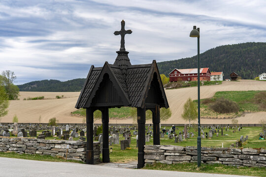 Traditional Norwegian stave church surrounded by spring countryside and forested hills under dramatic cloudy sky in Telemark, Norway. Dark wooden medieval-style architecture. 