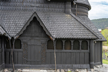 Obraz premium Traditional Norwegian stave church surrounded by spring countryside and forested hills under dramatic cloudy sky in Telemark, Norway. Dark wooden medieval-style architecture. 