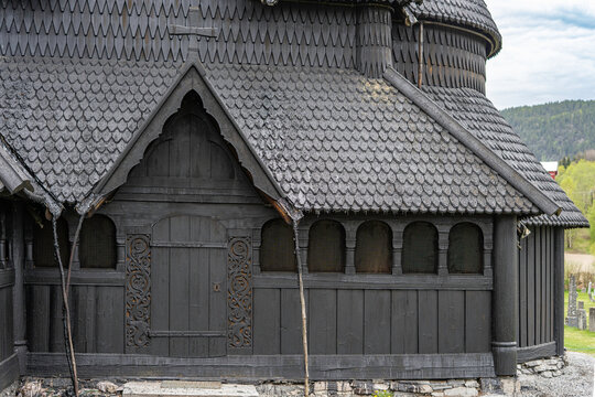 Traditional Norwegian stave church surrounded by spring countryside and forested hills under dramatic cloudy sky in Telemark, Norway. Dark wooden medieval-style architecture. 