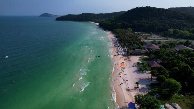 Aerial video of the amazing emerald water white sand Sao Beach with lively water sports, tourists, cabanas and sun umbrellas, beach bar and parasailing. Hot sunny day with long waves on Phu Quoc Islan