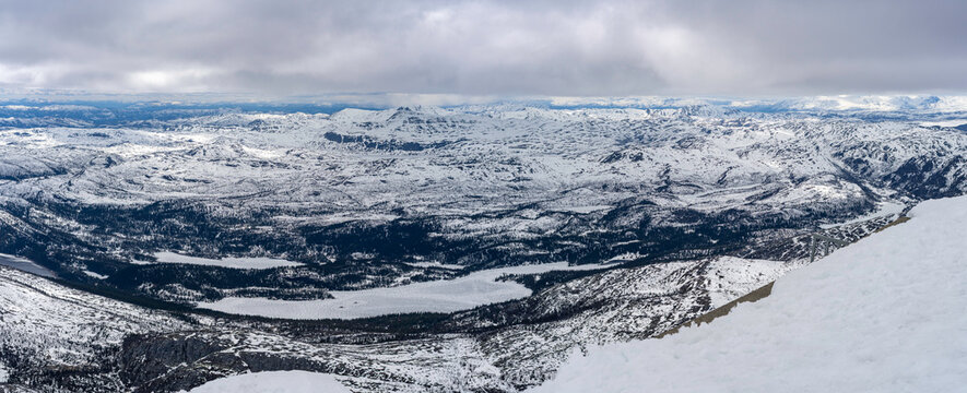 Panoramic view from the summit of Gaustatoppen at 1,850 meters above sea level. Snow-covered mountain plateaus, rocky ridges, and vast landscapes of southern Norway under a dramatic cloudy sky.