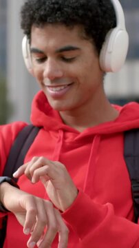 Vertical video: Tapping student in red hoodie checking smartwatch at city plaza wearing headphones