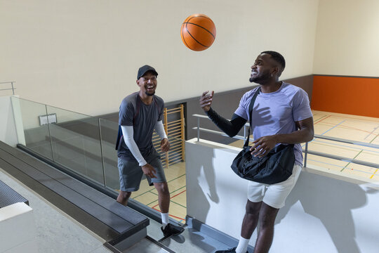 Diverse male friends tossing basketball and reaching on bleacher steps at gym with duffel bag