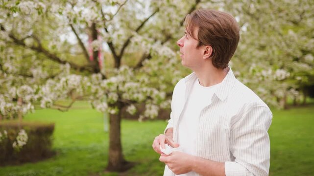 Man uses a tissue for hay fever symptoms during allergy season. 4K video showing the impact of pollen from spring blooms on itchy eyes and nose in a park. High quality footage