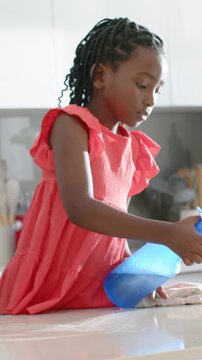 Vertical video: Grasping kid in red dress holding blue bottle and rag in kitchen adult guiding wipe