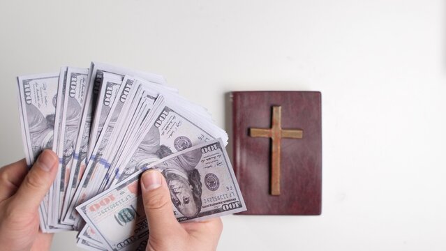 Hands fanning hundred dollar bills beside a wooden cross on a bible, top view studio shot symbolizing money, faith, tithing, greed, charity and moral conflict