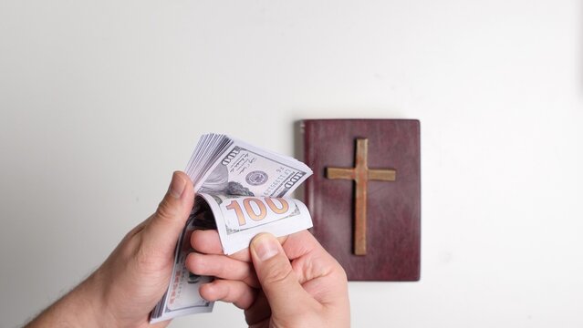 Hand holding and counting a stack of hundred dollar banknotes near a book with a cross, symbolizing themes of religion, money, tithe, corruption, and financial offerings